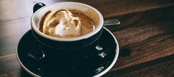A cup of coffee topped with whipped cream on a black saucer, resting on a dark wooden table.